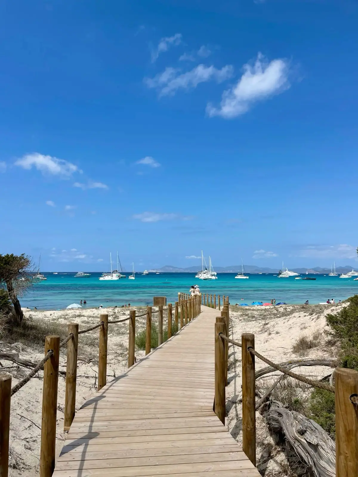 Pasarela de madera hacia una playa de Formentera, con arena, aguas turquesas y barcos al fondo.